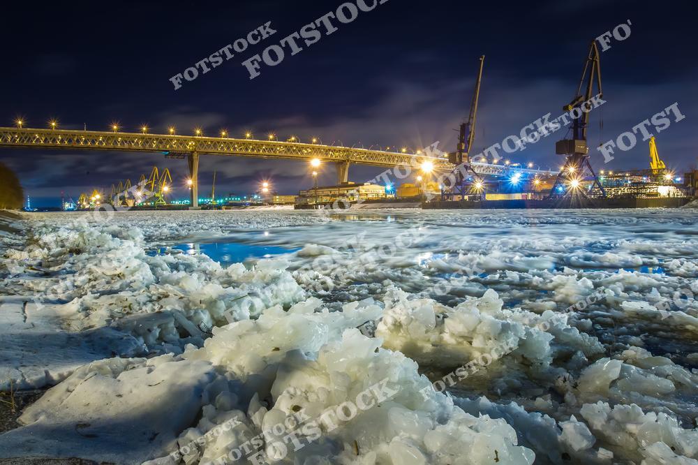 На фотографии изображён замерзший участок реки в ночное время. Виден ярко освещённый мост, предположительно относящийся к индустриальной зоне Санкт-Петербурга. На заднем плане можно увидеть портовые краны, работающие в ночной подсветке. Зимняя атмосфера подчёркивается льдом и снегом на переднем плане.